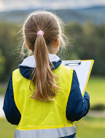 A girl overlooks a green space, she is wearing a high vis jacket and holding a clipboard.